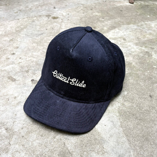 Blue cap with 'Critical Slide' embroidery on a concrete surface