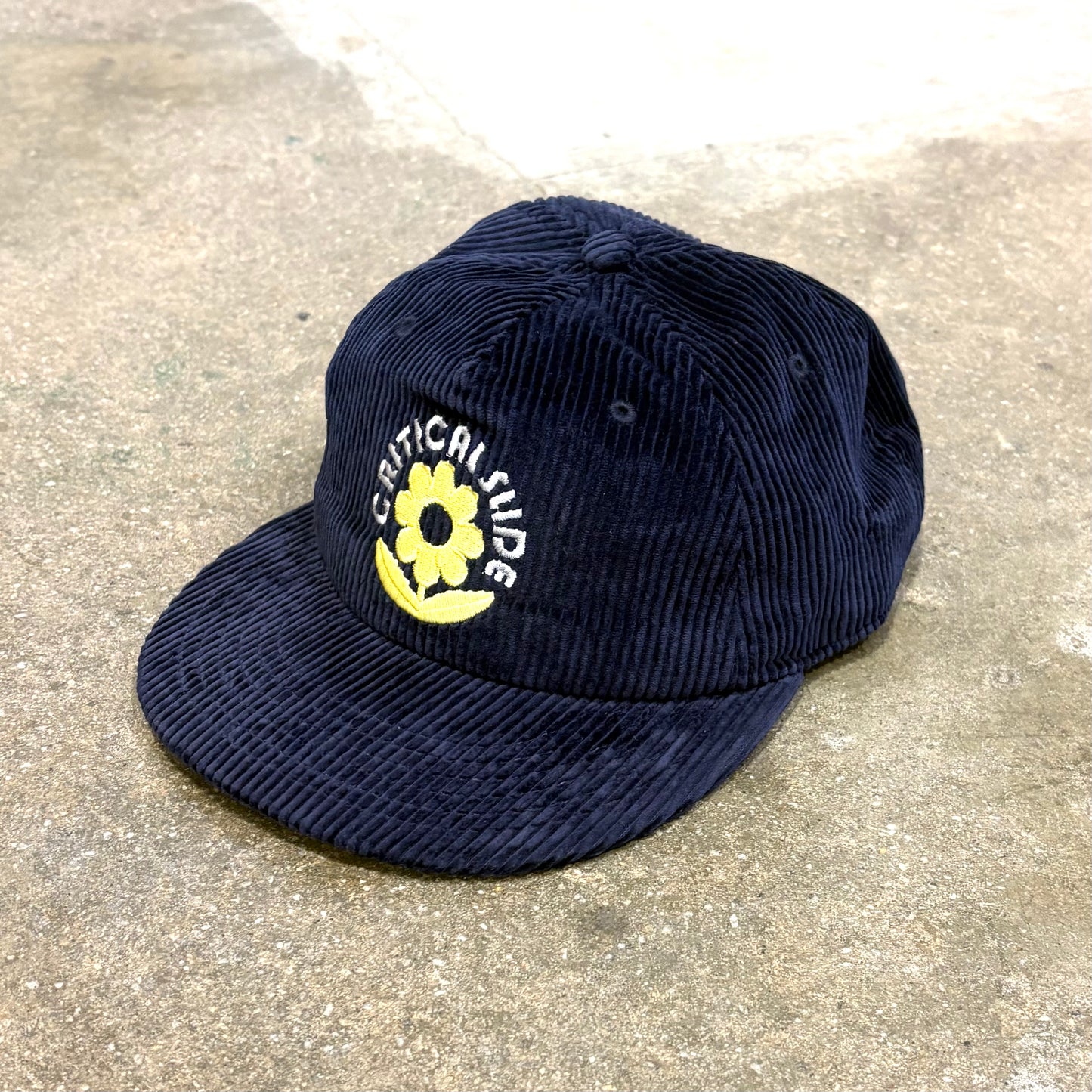 Navy blue corduroy cap with a yellow flower and white text on a concrete surface