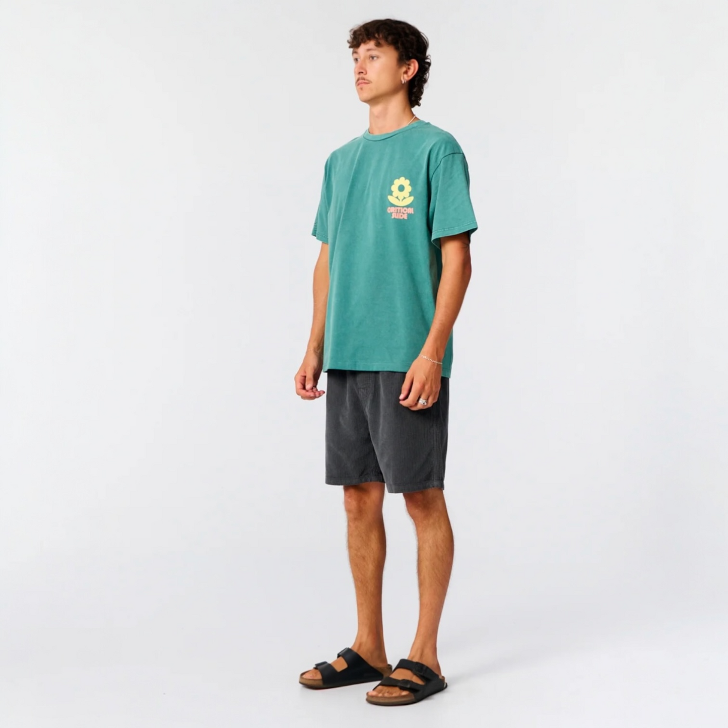 Person wearing a teal t-shirt with a logo on the chest and gray shorts against a white background