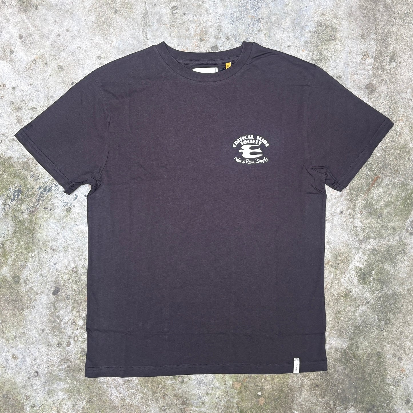 A black short-sleeve crewneck t-shirt with a small white graphic on the left chest, laid flat on a textured concrete surface.