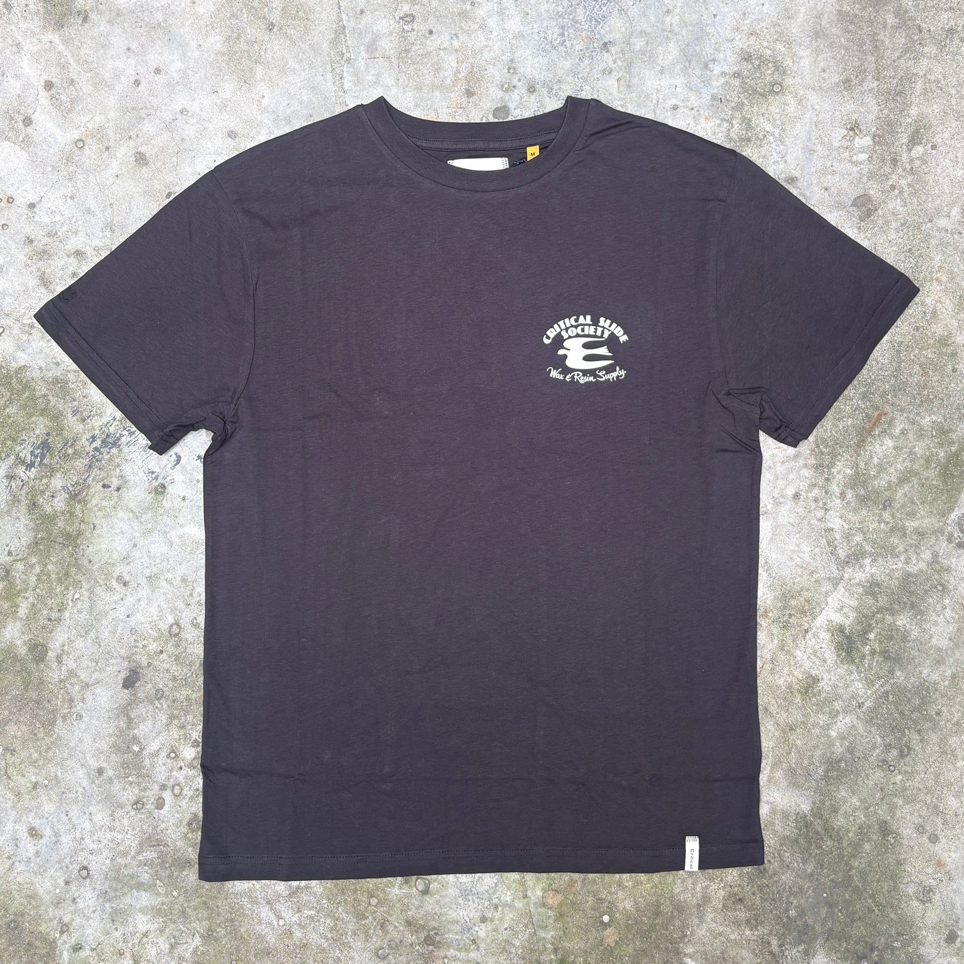 A black short-sleeve crewneck t-shirt with a small white graphic on the left chest, laid flat on a textured concrete surface.
