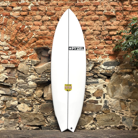 White Pyzel Tiger Twin surfboard with a brand logo against a rustic stone wall.