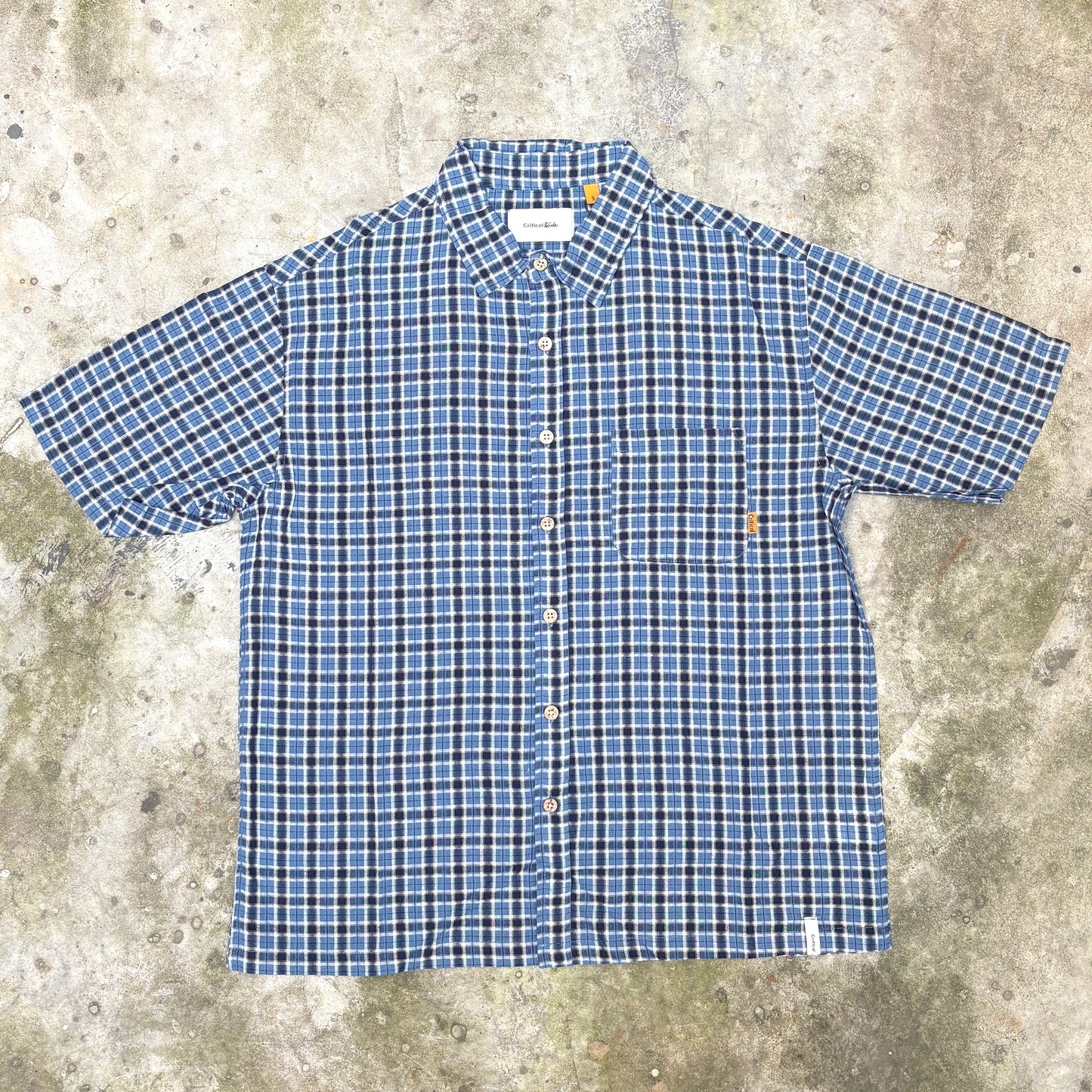 A short-sleeve button-up shirt with a blue, white, and black plaid pattern, laid flat on a textured concrete surface.
