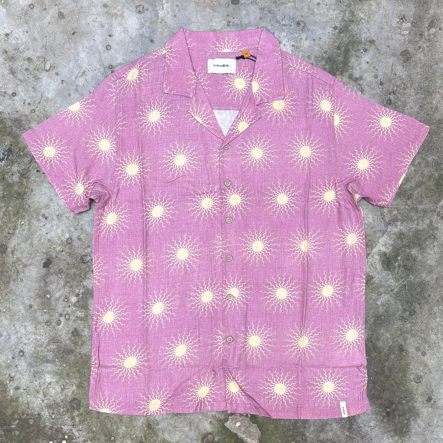 A short-sleeve button-up camp shirt in pink with a repeating cream sunburst pattern, laid flat on a textured gray concrete surface.
