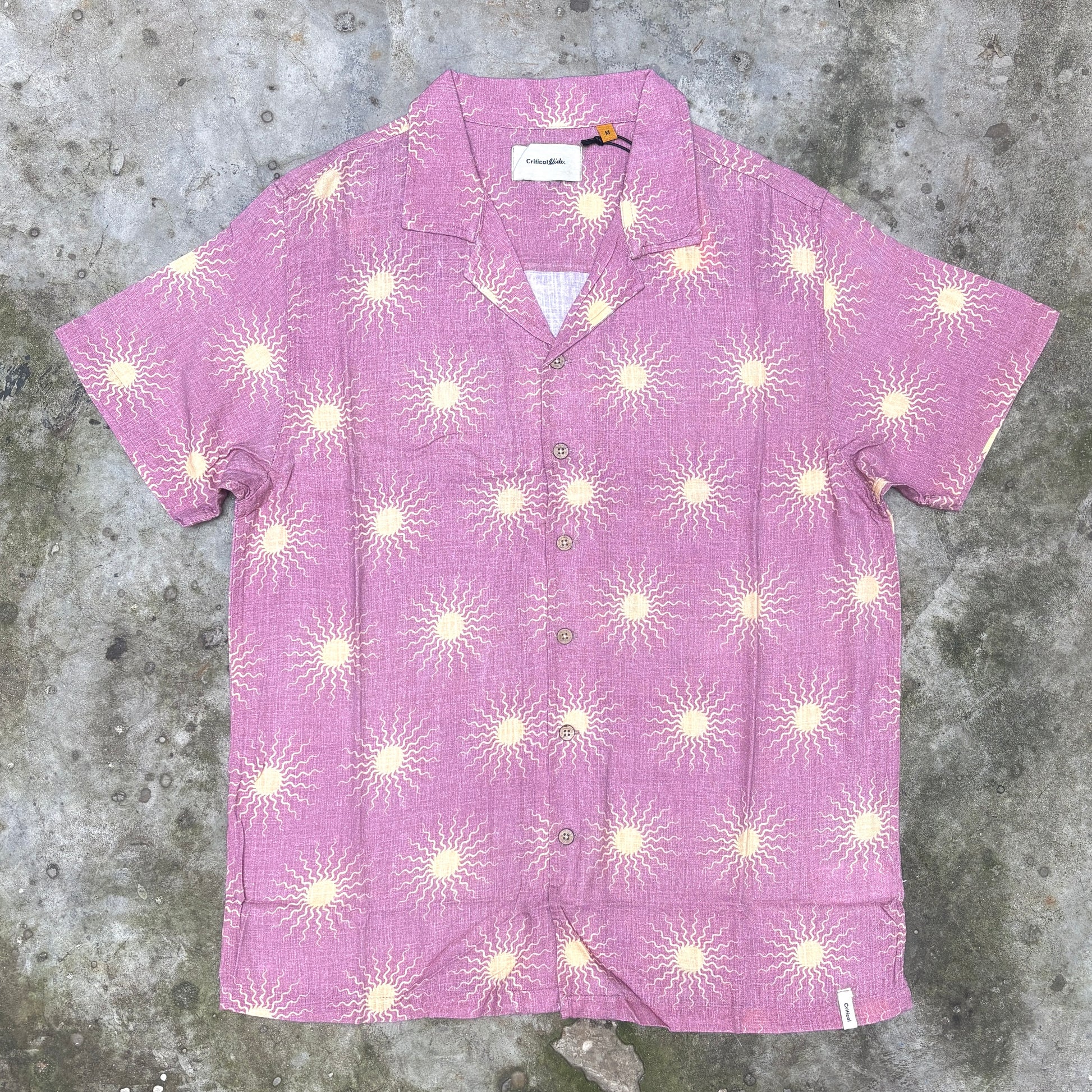 A short-sleeve button-up camp shirt in pink with a repeating cream sunburst pattern, laid flat on a textured gray concrete surface.