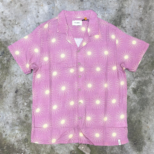 A short-sleeve button-up camp shirt in pink with a repeating cream sunburst pattern, laid flat on a textured gray concrete surface.