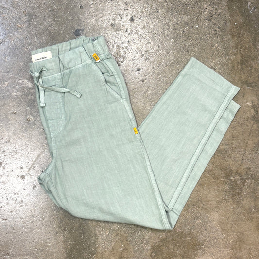 A pair of sage green linen drawstring pants laid flat on a textured concrete floor. The pants feature a relaxed, straight-leg fit, a drawstring waist, and small yellow brand tags.