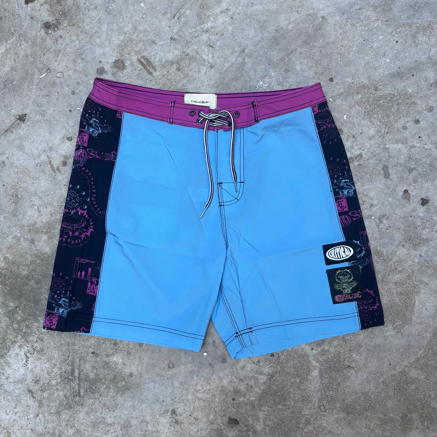 Blue shorts with purple waistband and graphic sides on a concrete surface