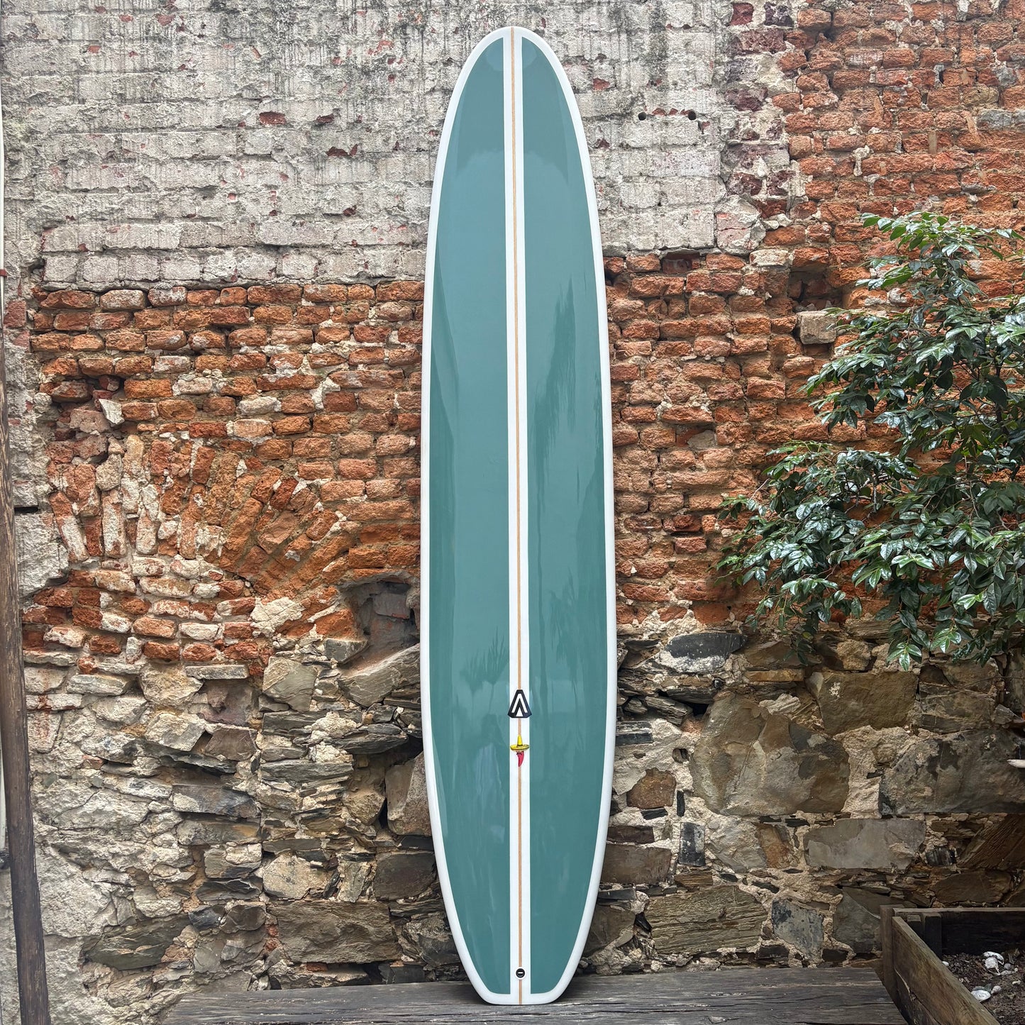 Teal longboard with a white logo against a rustic stone wall.