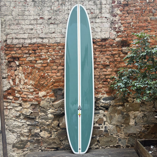 Teal longboard with a white logo against a rustic stone wall.