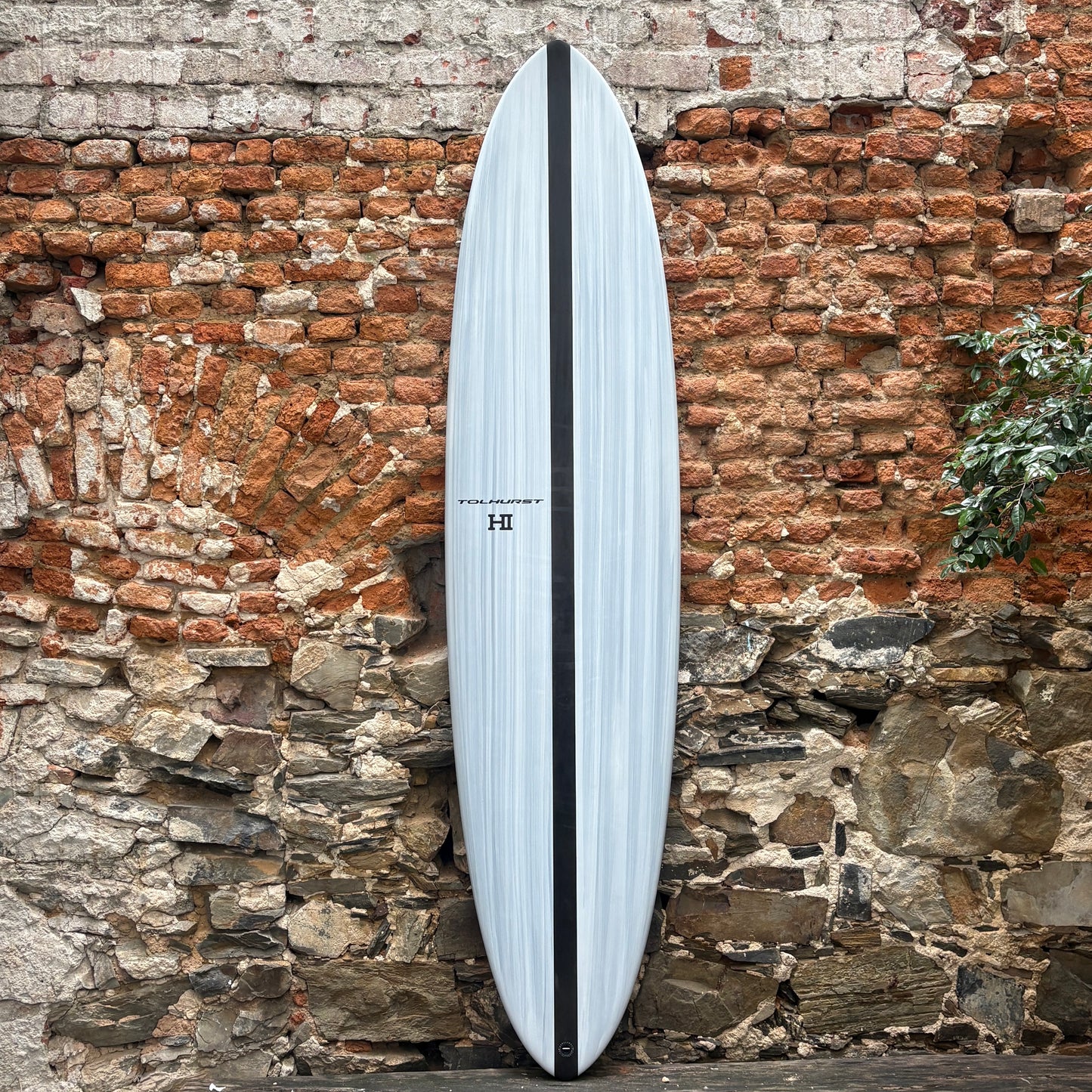 A long surfboard with a pale blue-gray body, black center stripe, and small HI branding, leaning against a rustic brick and stone wall.