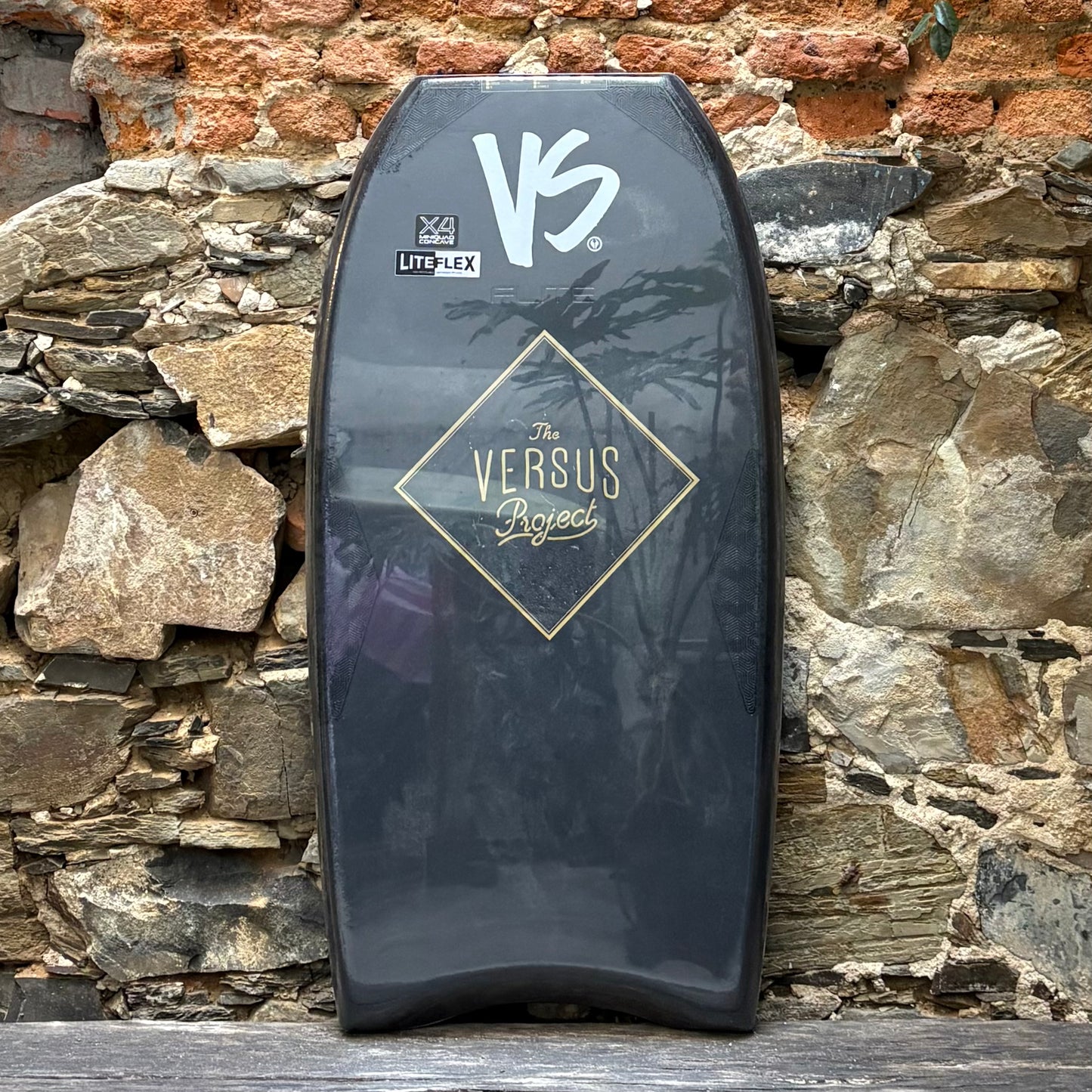 Black bodyboard with 'Versus' branding against a stone wall background