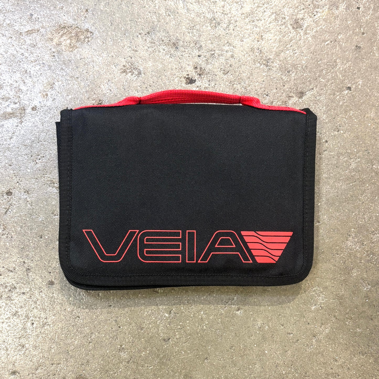 A black zippered pouch or organizer with a red top handle and a red outlined VEIA logo on the front, photographed on a gray concrete surface.