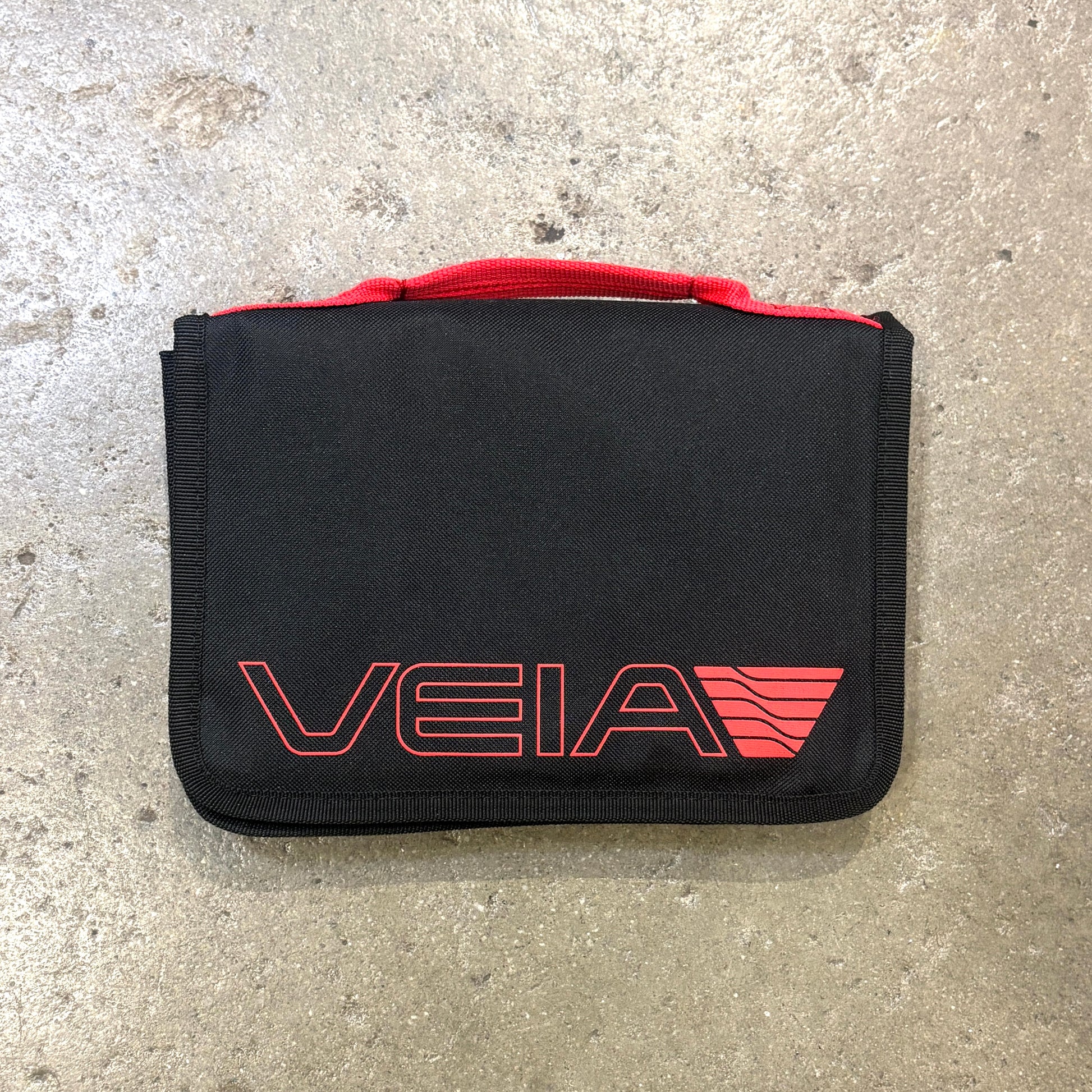 A black zippered pouch or organizer with a red top handle and a red outlined VEIA logo on the front, photographed on a gray concrete surface.