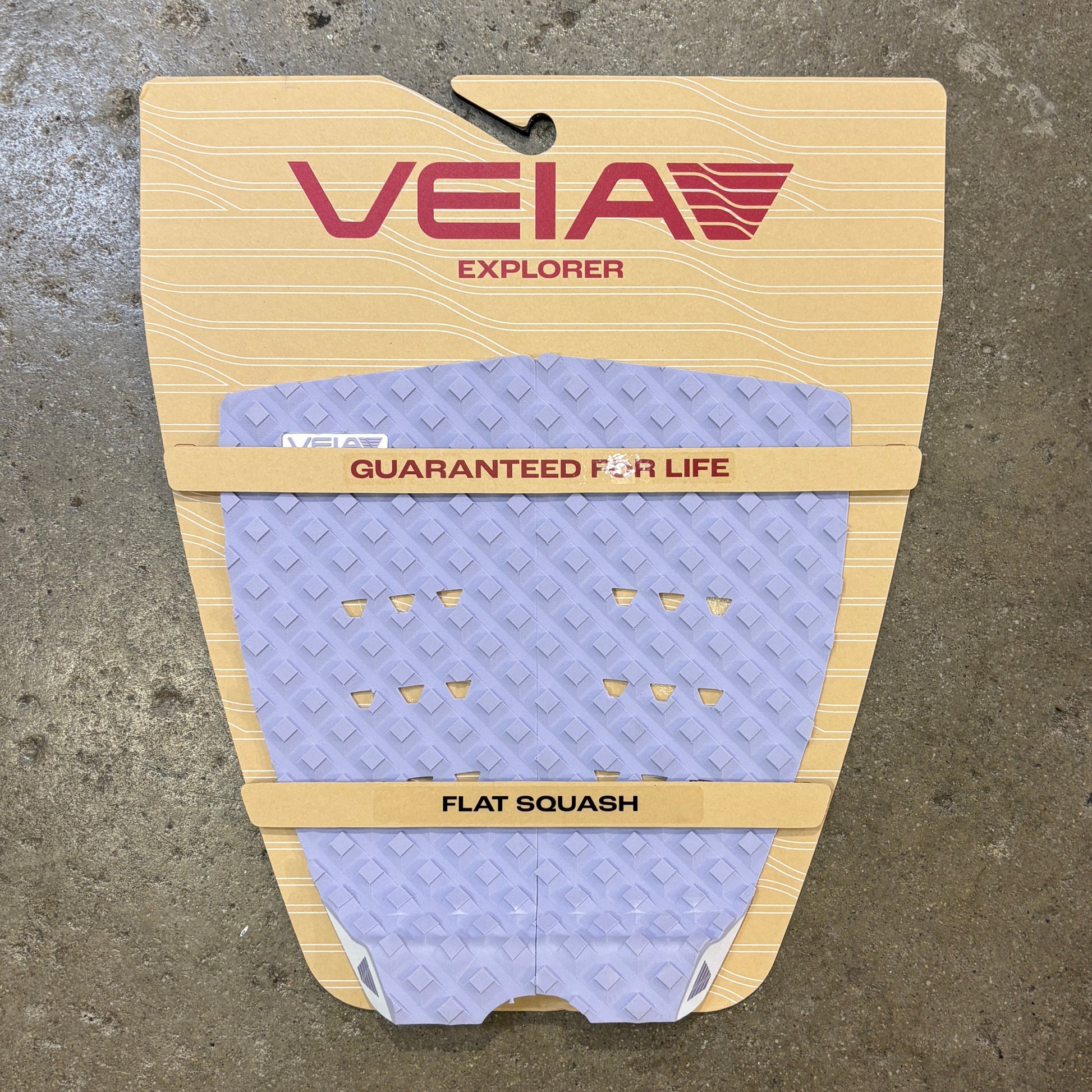 A Veia Explorer flat squash surfboard grip pad in lavender purple with a diamond pattern, mounted on its beige cardboard backing against a concrete floor.