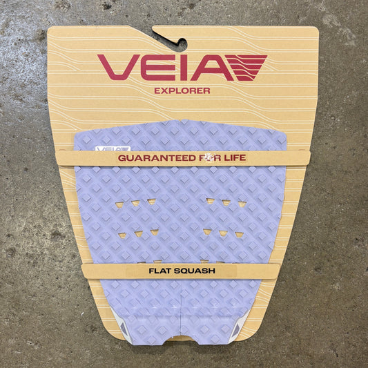 A Veia Explorer flat squash surfboard grip pad in lavender purple with a diamond pattern, mounted on its beige cardboard backing against a concrete floor.