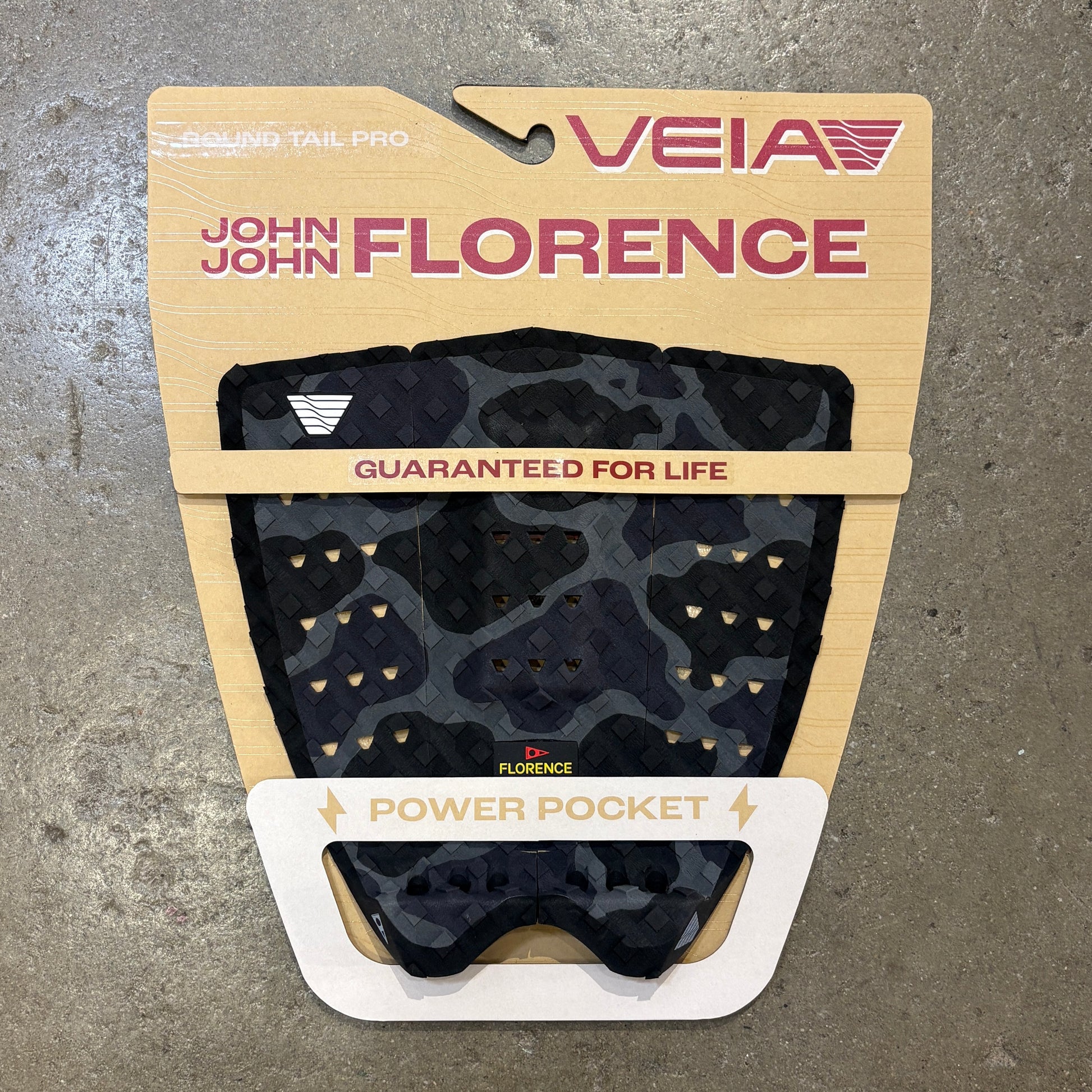 Veia John John Florence Power Pocket tailgate pad in black and dark gray camouflage, mounted on its original beige cardboard backing against a gray concrete floor.