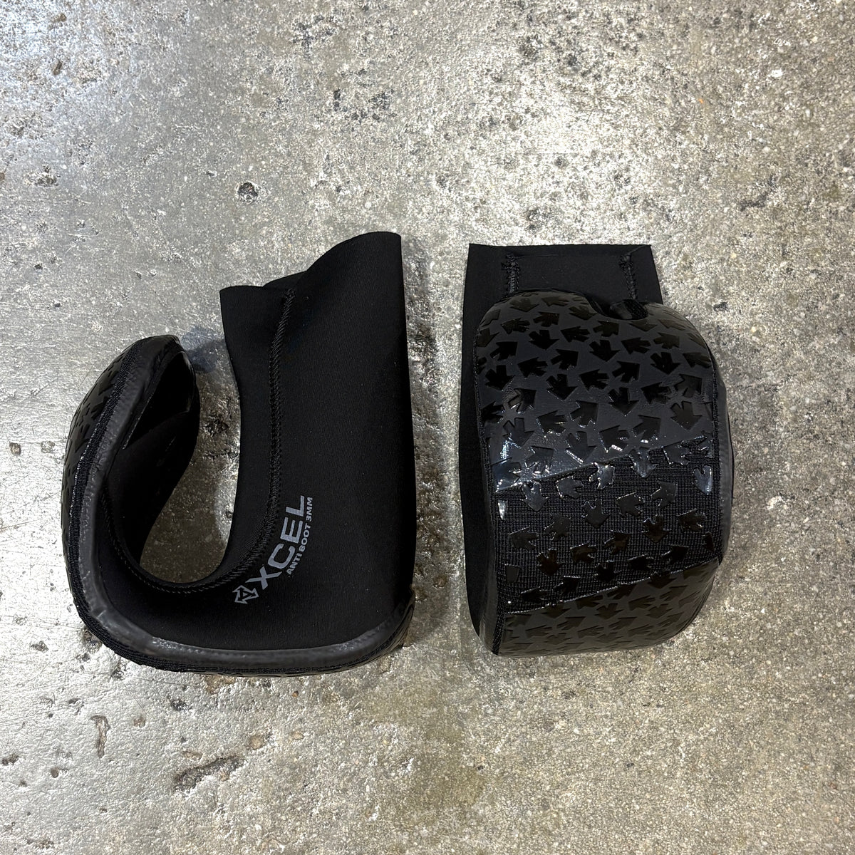 A pair of black neoprene and carbon-fiber wing warmer boots for snowboarders, displayed side-by-side on a textured concrete floor. One boot is shown closed, revealing the black interior and a white 'XCEL' logo, while the other is shown open to display the black carbon-fiber patterned exterior.