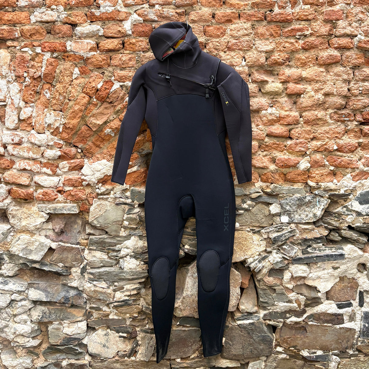 A black full-body wetsuit with a hood and knee pads, hanging against a rustic brick and stone wall.