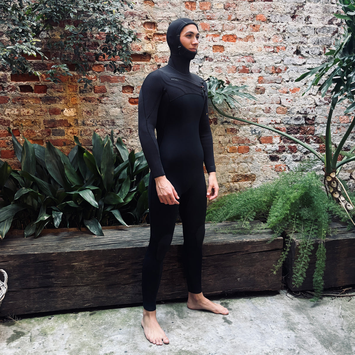 A man wearing a full-body black wetsuit with a hood stands barefoot outdoors against a rustic brick wall with green plants.