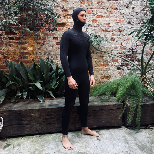 A man wearing a full-body black wetsuit with a hood stands barefoot outdoors against a rustic brick wall with green plants.