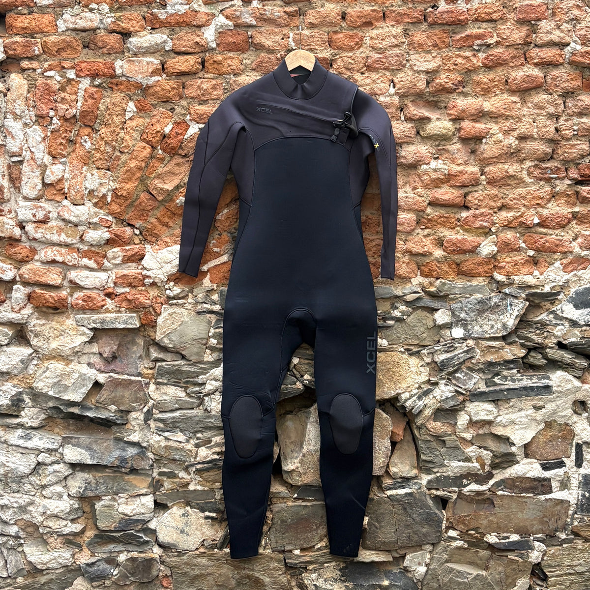 A black full-body wetsuit with black shoulder panels hangs on a wooden hanger against a rustic brick and stone wall background.