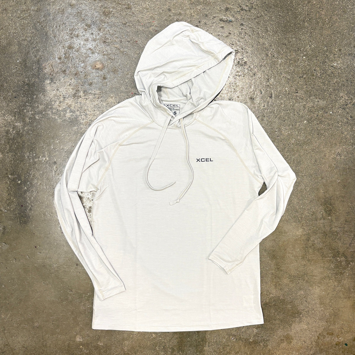 A white long-sleeve hooded athletic shirt with a small black XCEL logo on the left chest, laid flat on a textured concrete surface.