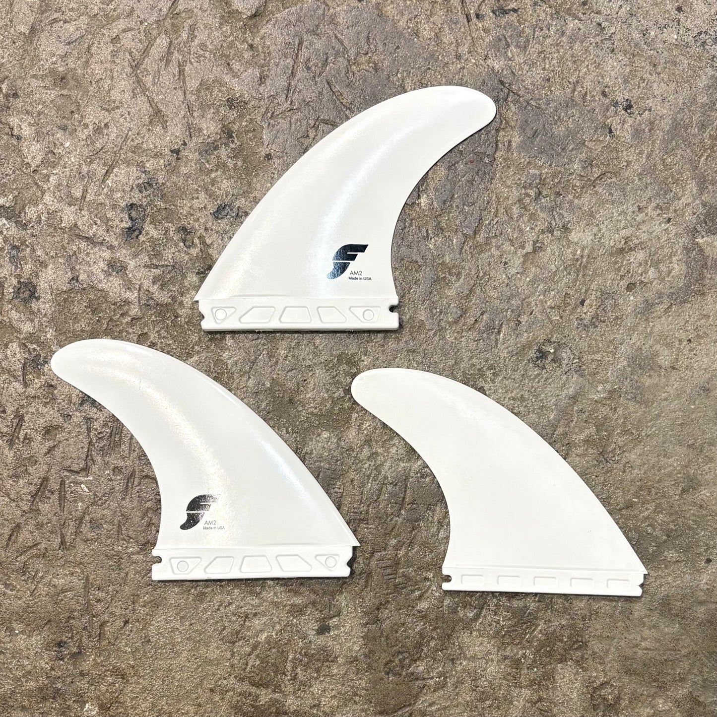 Three white surfboard fins with a logo on a textured brown surface