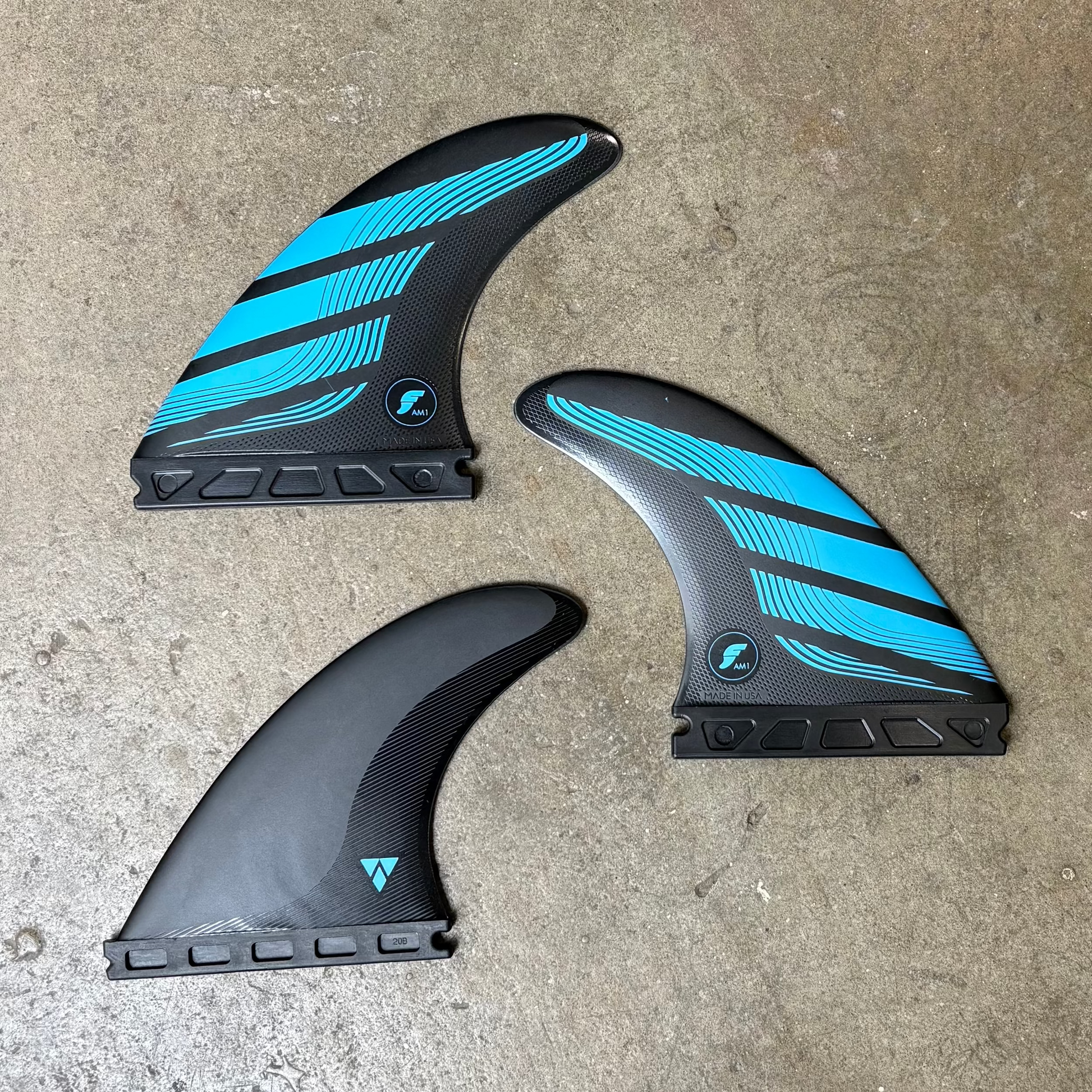 Three black and blue futures AM1 surfboard fins on a concrete surface