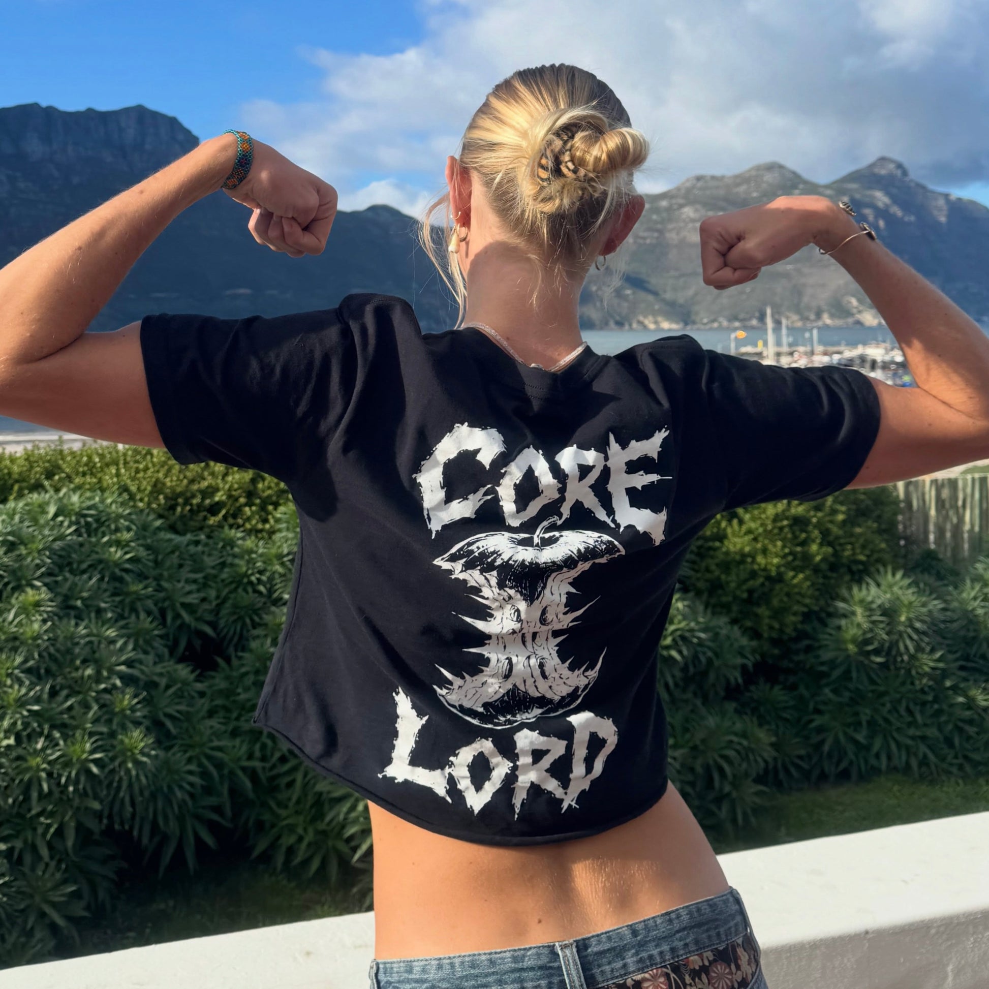 A woman viewed from behind wearing a black cropped t-shirt with a distressed white graphic that reads 'CORE A LORD' on the back, posing with her arms raised in front of a scenic outdoor background of mountains and greenery.