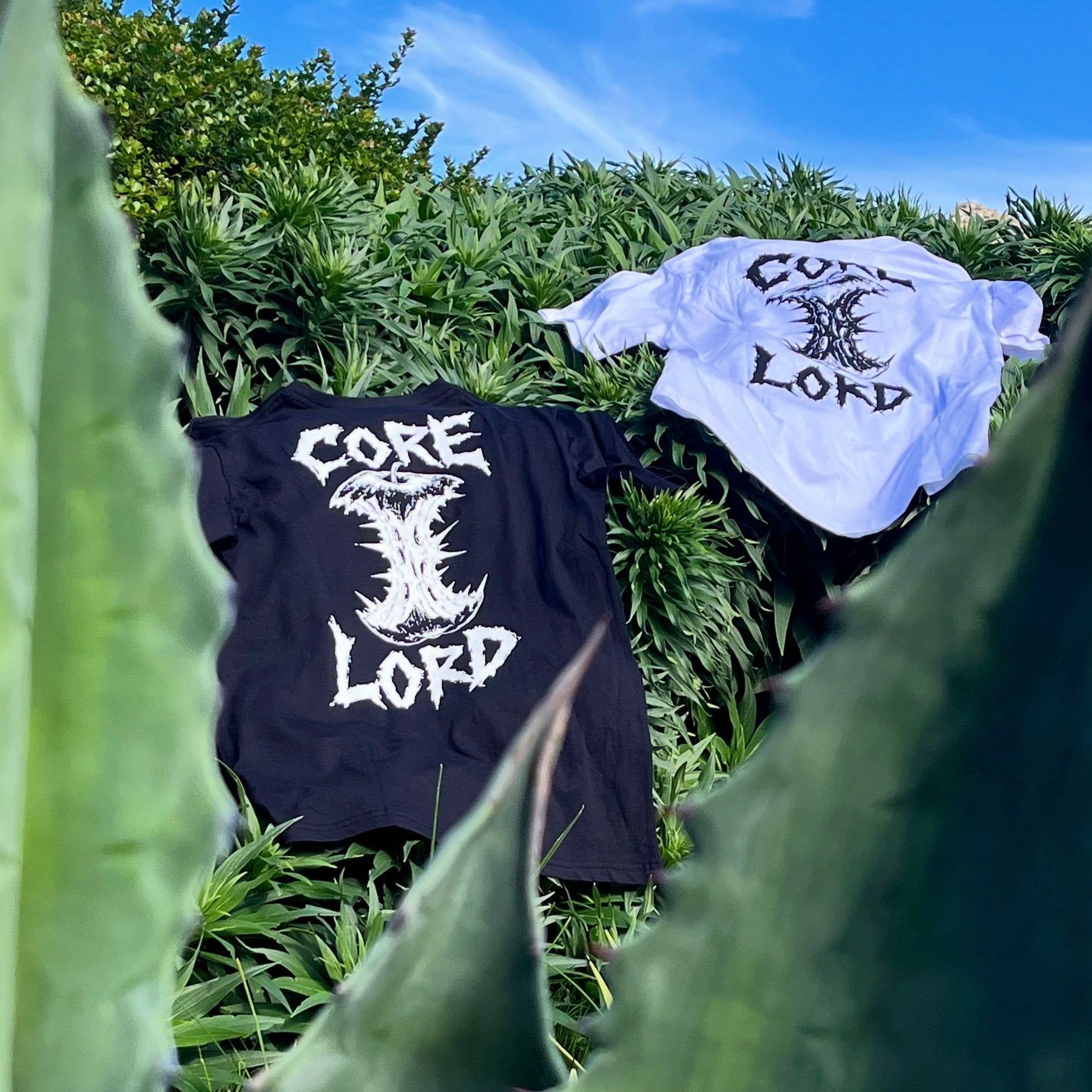 Two graphic t-shirts displayed outdoors on green foliage. One black t-shirt on the left features a white gothic-style mushroom graphic with the text 'CORE LORD'.