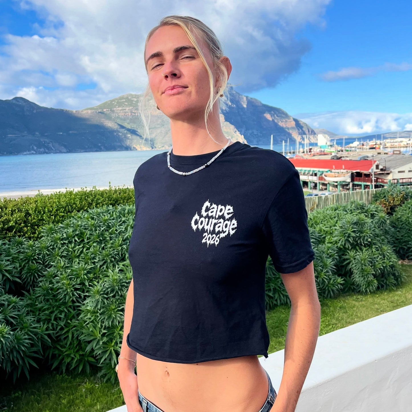 A woman wearing a black cropped t-shirt with 'Cape Courage 2025' printed in white on the chest, standing outdoors with a scenic coastal background.