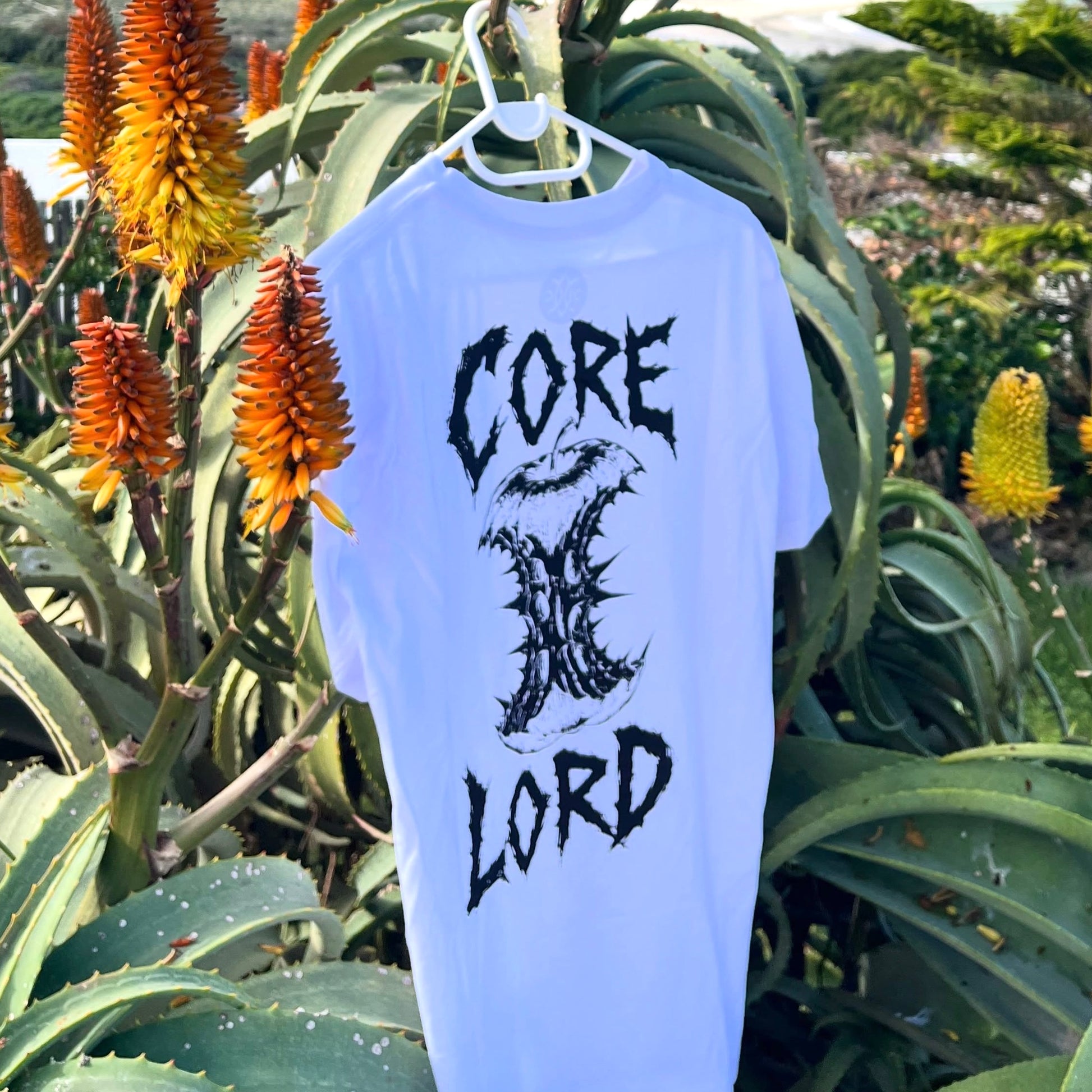 A light blue t-shirt hanging on a white plastic hanger, featuring a black graphic print that reads 'CORE X LORD' with a stylized X logo. The shirt is photographed outdoors against a background of green aloe vera leaves and orange flowers.