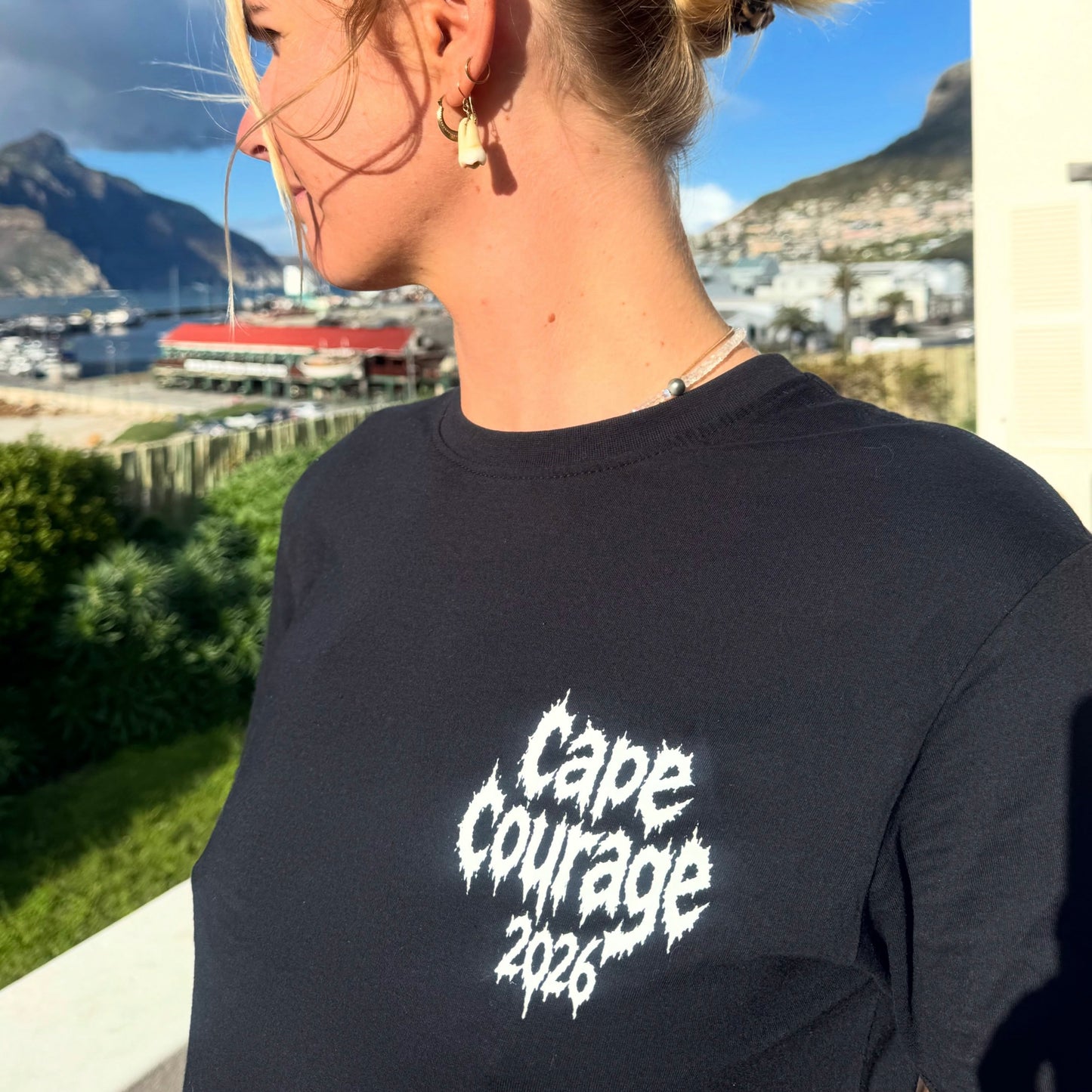 A woman wearing a black t-shirt with a white distressed graphic that reads 'Cape Courage 2026', photographed outdoors with a scenic coastal background.