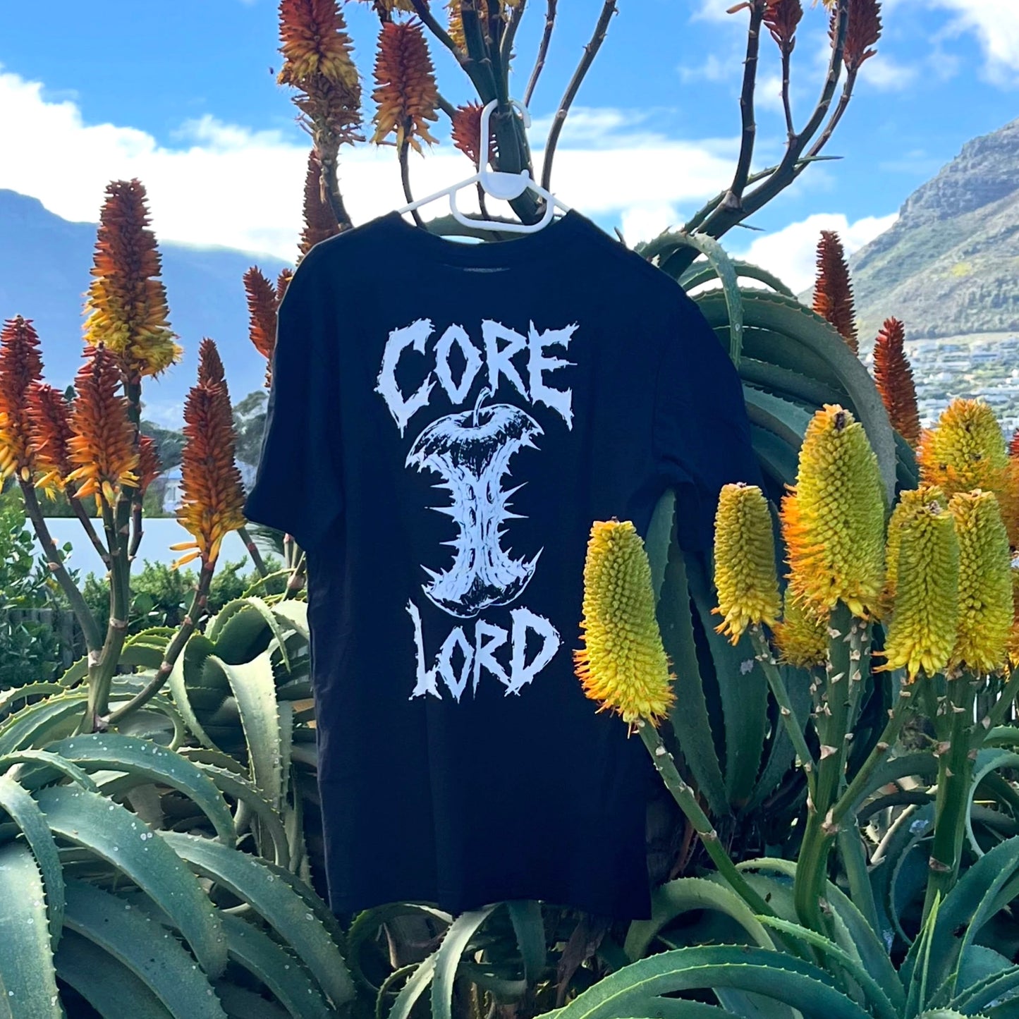 A black short-sleeve graphic T-shirt hanging outdoors on a hanger, featuring a large white stylized 'CORE LORD' text and skull design on the front. The shirt is surrounded by green aloe plants and orange-yellow flowers, with mountains and blue sky in the background.