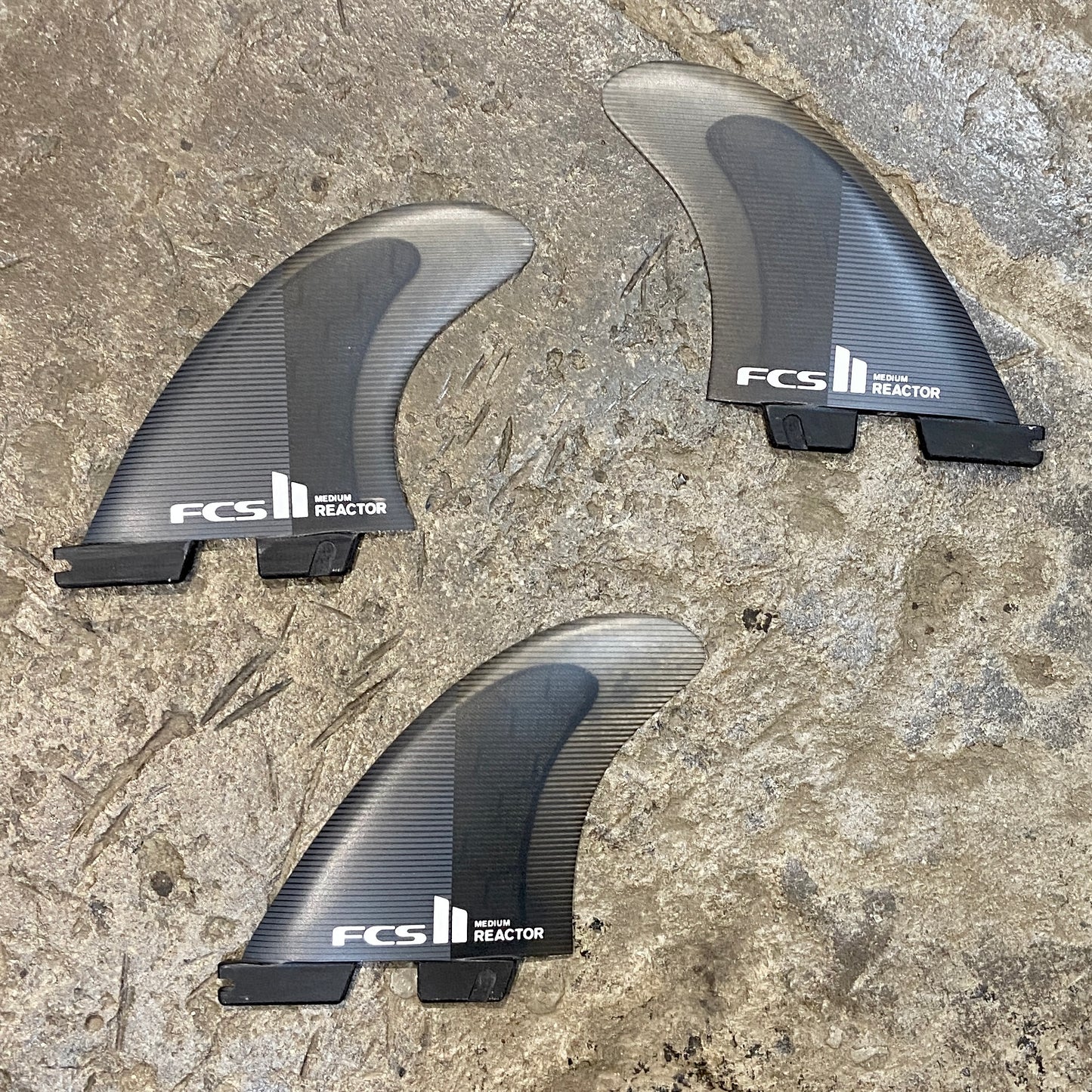 A medium set of FCS II Reactor PC Tri Fins in a black and grey colour way laid on a concrete surface.