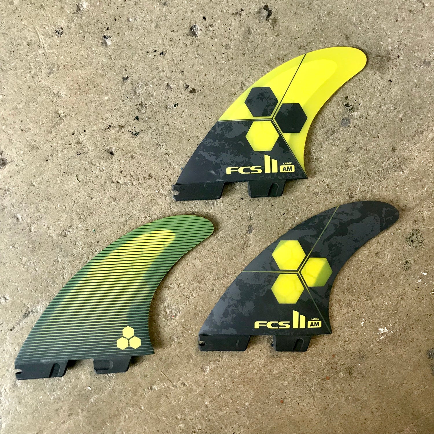 A large set of the FCS II Shaper Series Al Merrick Tri Fin in a black and yellow colour way laid out on a concrete surface