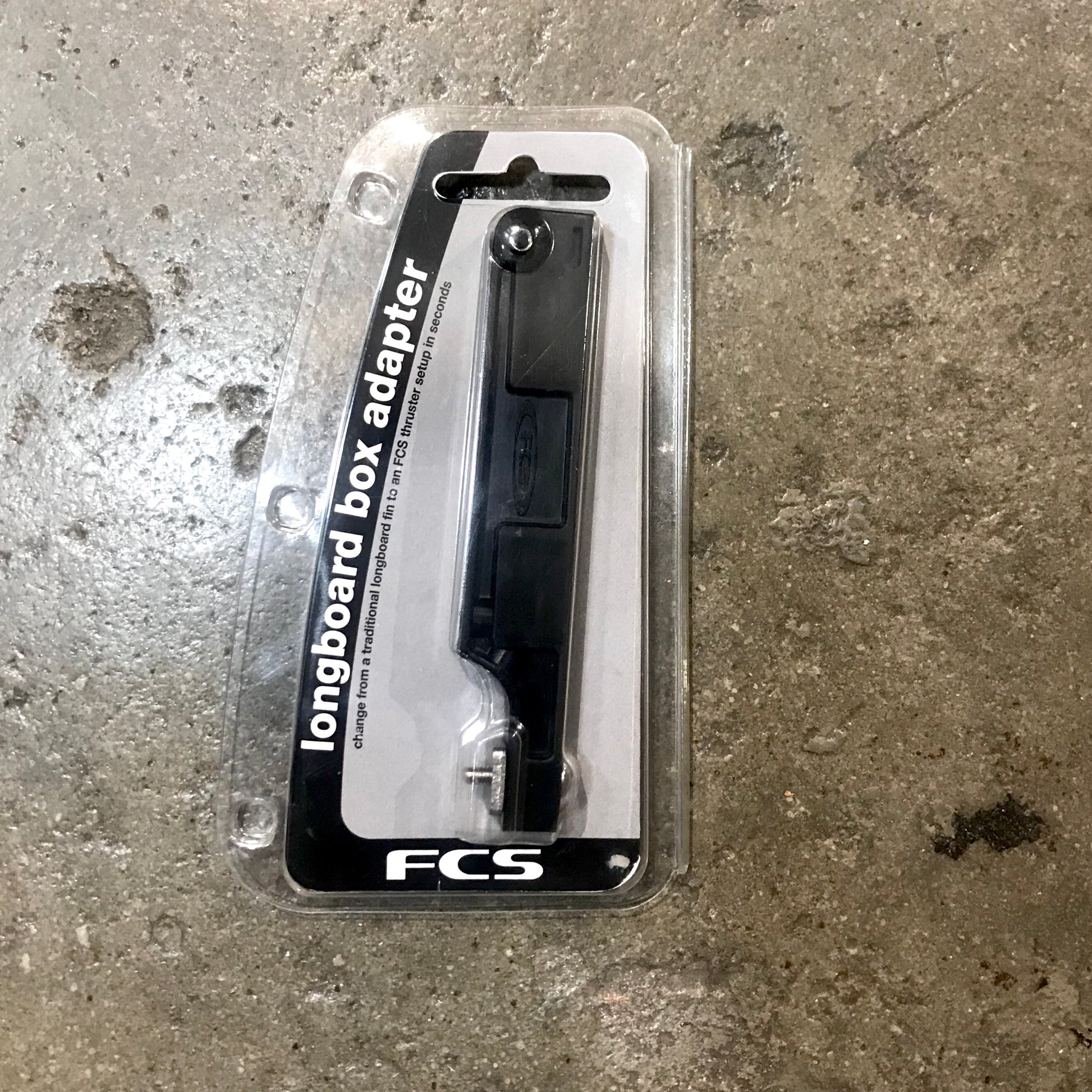 A FCS Longboard Box Adapter in it's packaging laid out on a concrete surface