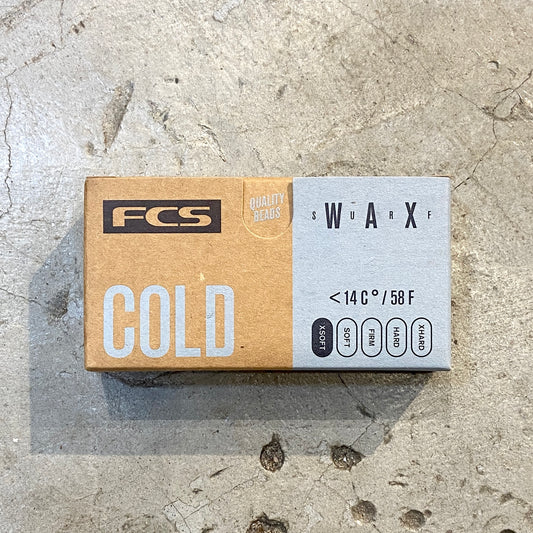A block of FCS Surf Wax Cold in brown and blue packaging laid on a concrete floor