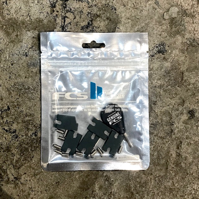 A packaged FCS tab infill kit containing several black plastic components, screws and a key, displayed on a textured concrete surface.