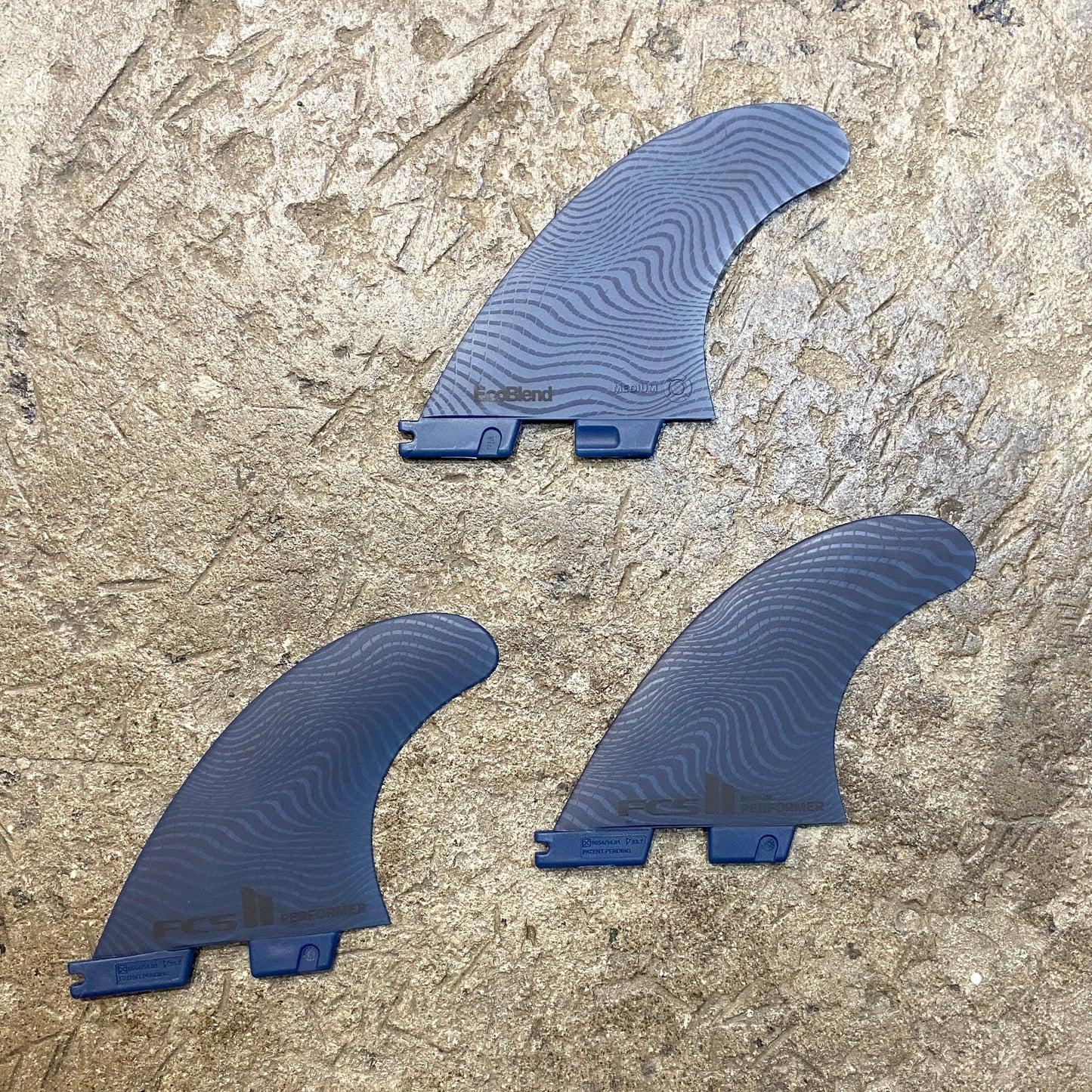 View of the FCS ll Performer Neo Glass Eco Tri Fins L in a blue colour way laid out on a concrete floor.