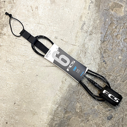 A FCS Competition Essential Leash 6ft/ 5.5mm in black laid out on a concrete surface
