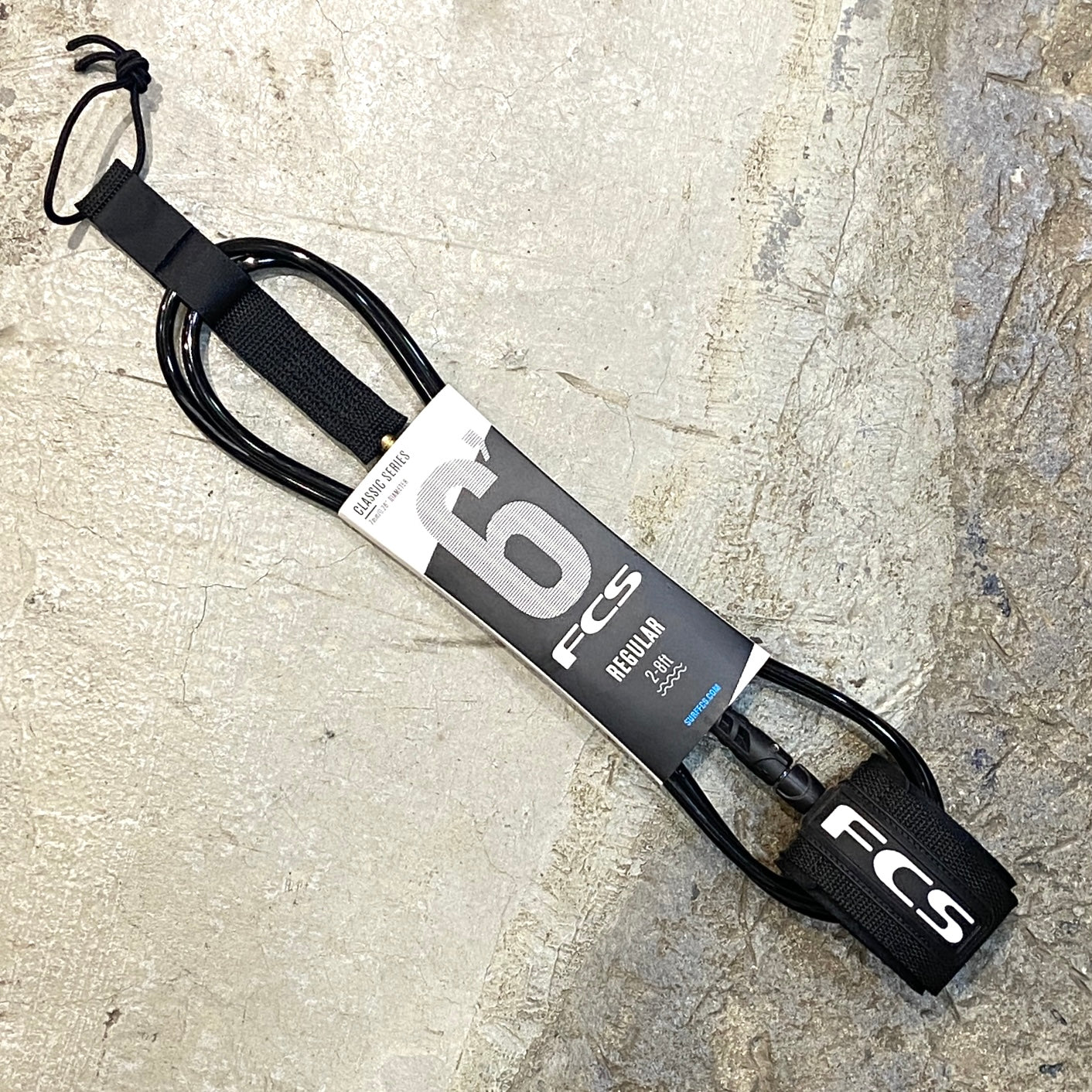 A FCS Regular Leash 6ft / 7mm in black laid out on a concrete floor