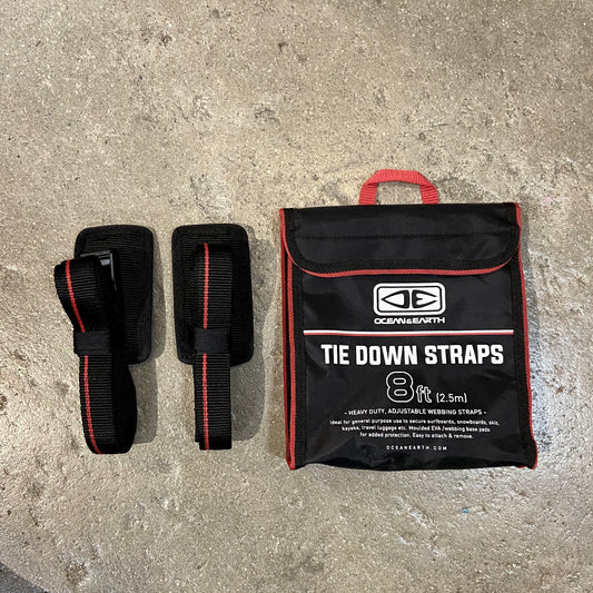 A set of black and red OCEAN&EARTH tie down straps with a protective rubber cam buckle, alongside a storage bag on a concrete surface.