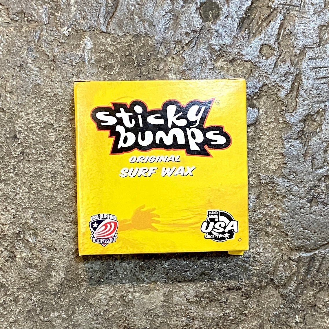 A yellow surf wax package labeled 'Sticky Bumps Original Surf Wax' with a graphic of a surfer on the front, indicating it's a product for tropical surf conditions.
