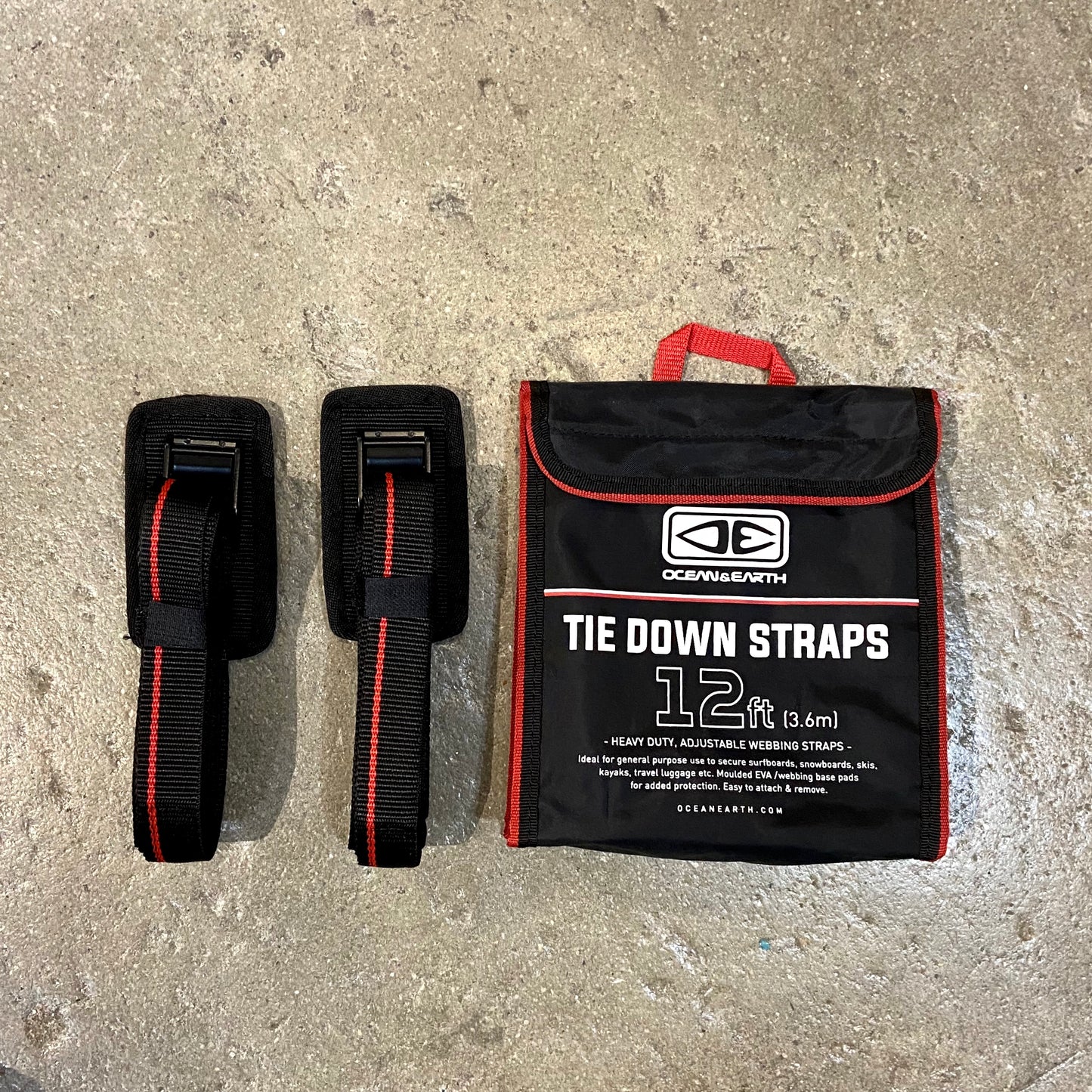 A set of black and red OCEAN&EARTH tie down straps with a protective rubber cam buckle, alongside a storage bag with the product information visible.